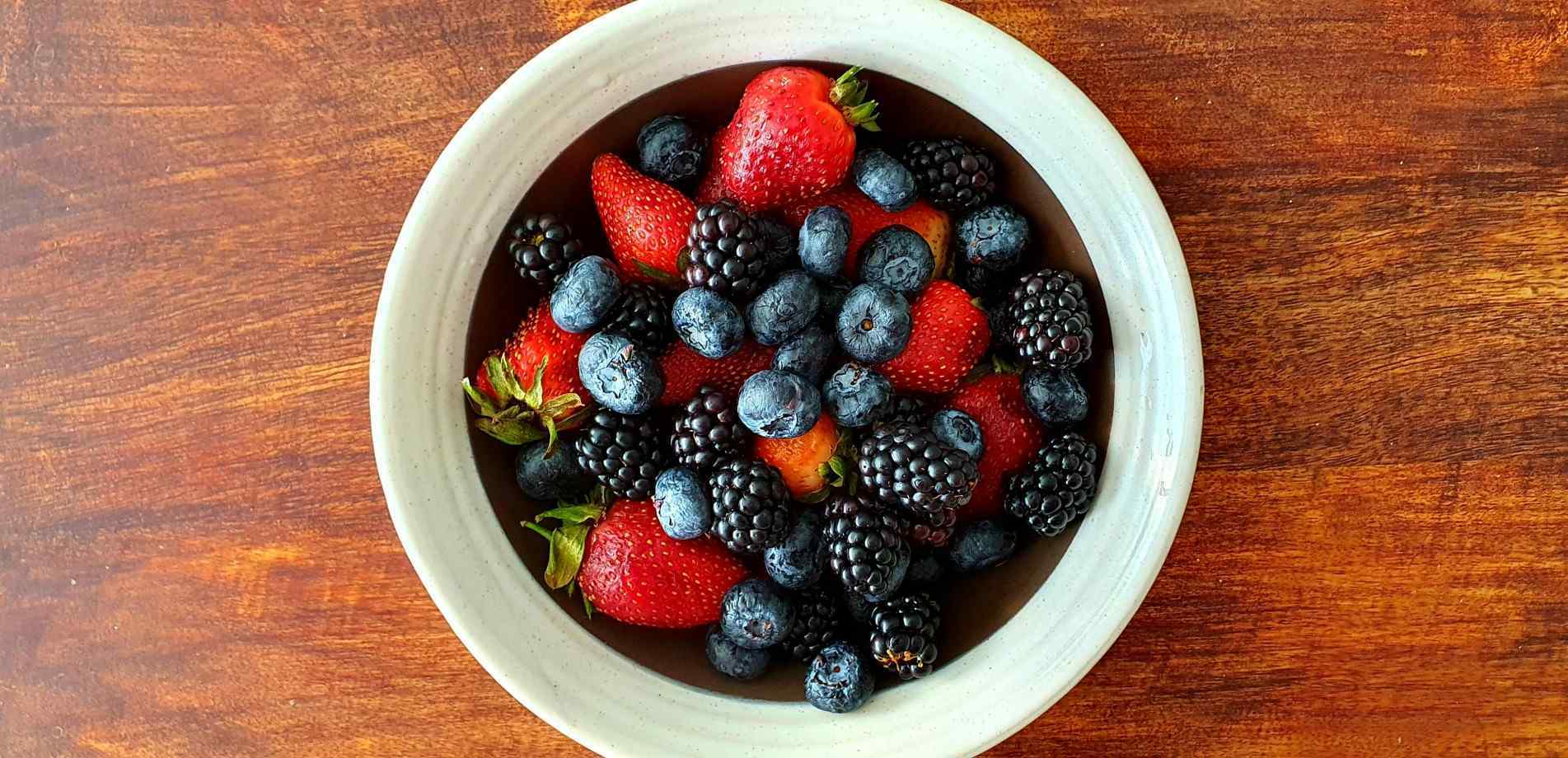 Tigela branca cheia de frutas vermelhas frescas, incluindo morangos, mirtilos e amoras, posicionada sobre uma mesa de madeira rústica
