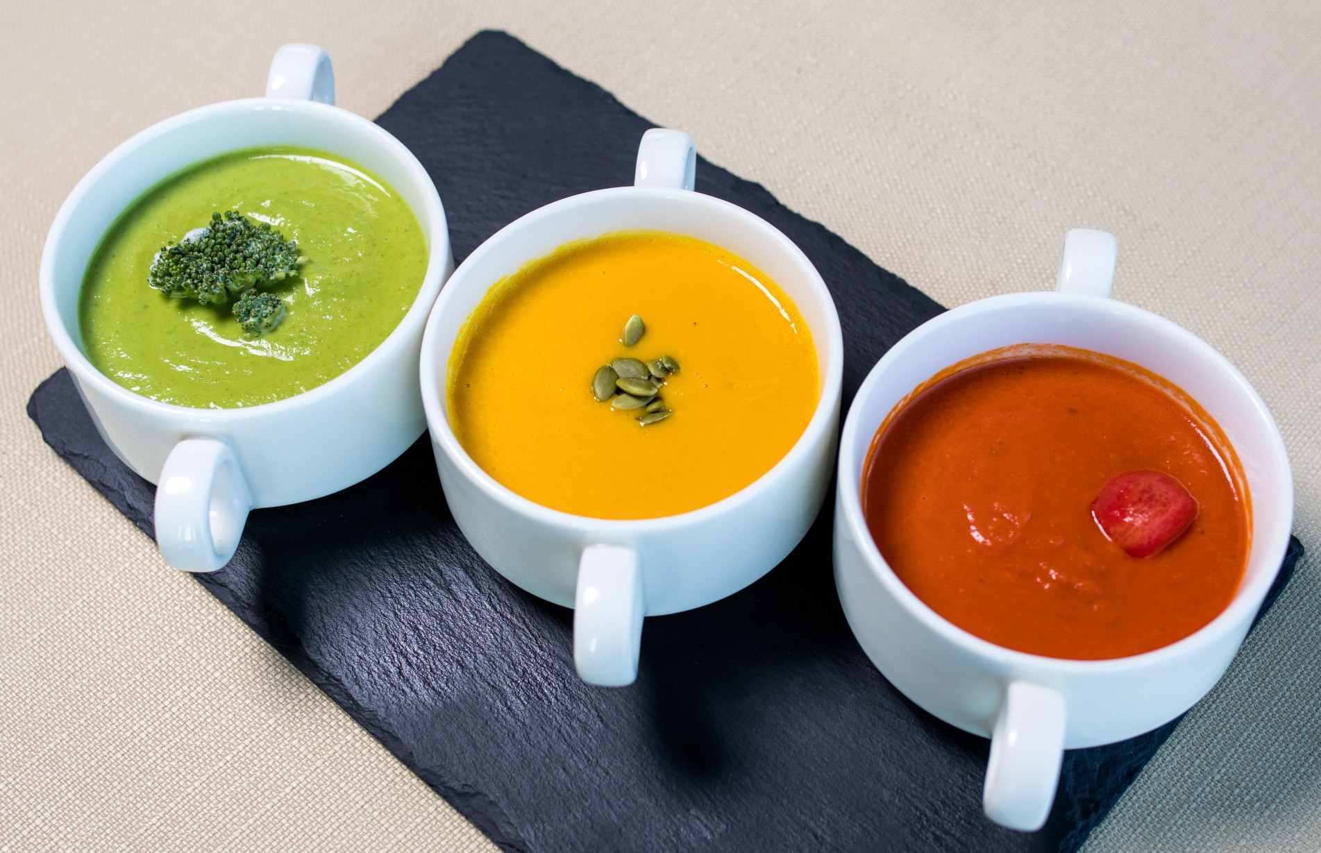Três tigelas brancas com sopas cremosas verde, amarela e vermelha servidas sobre uma tábua preta.
