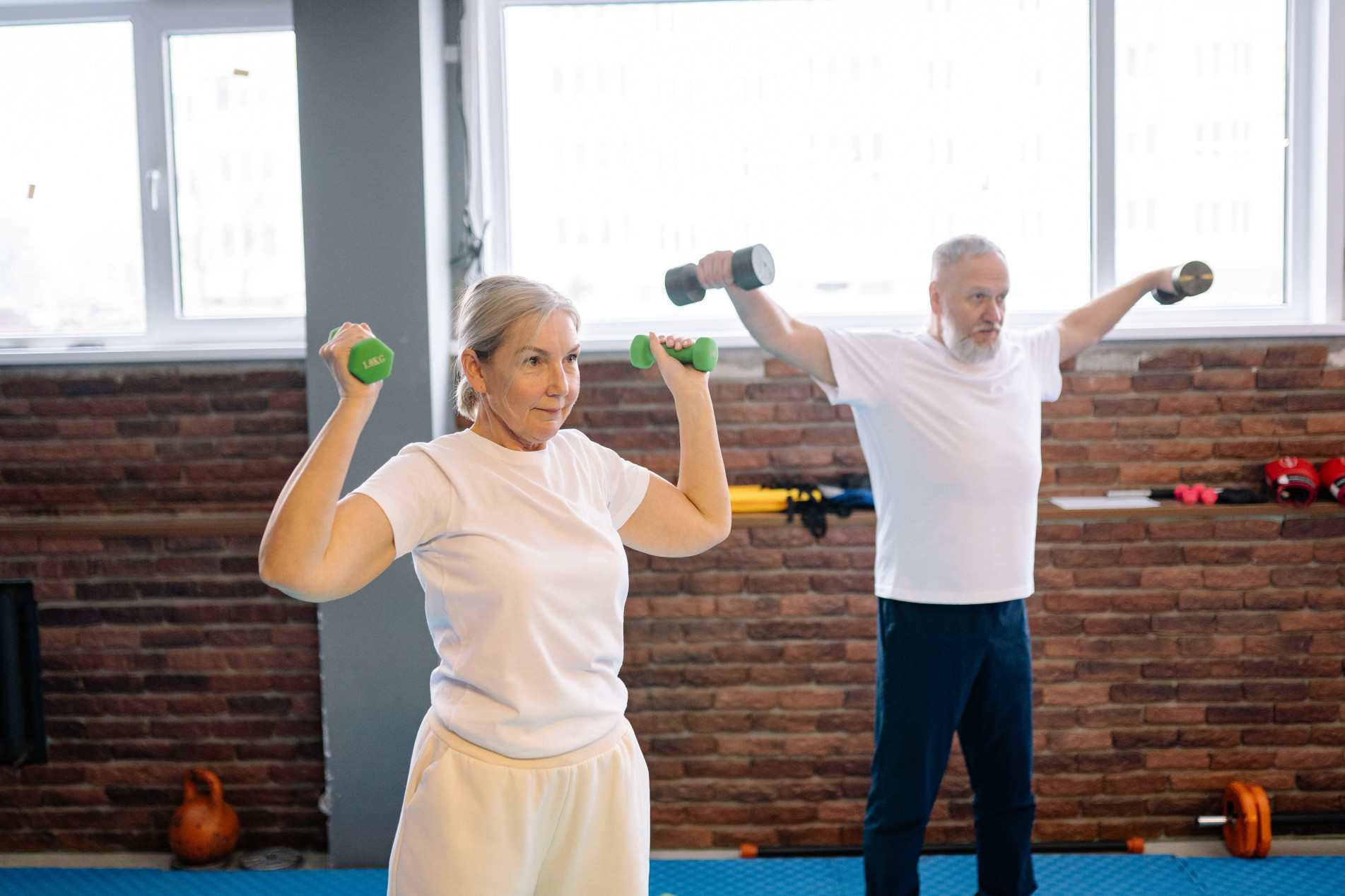 Um casal de idosos pratica exercícios de musculação levantando halteres dentro de uma academia bem iluminada.