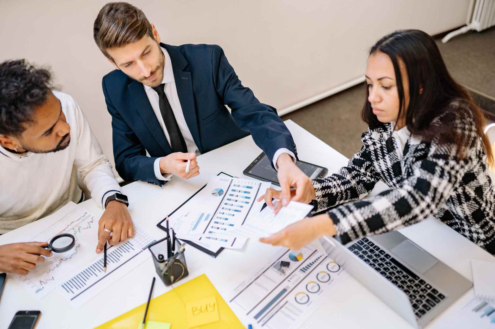 Três profissionais em uma reunião de negócios analisam e compartilham relatórios com gráficos e planilhas financeiras sobre uma mesa de escritório.