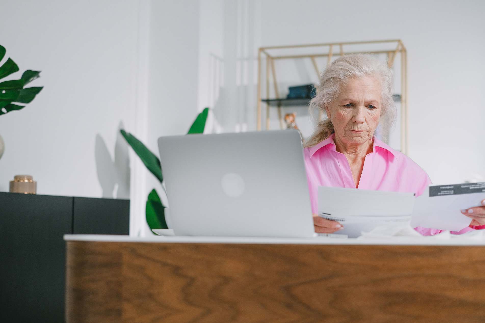 Mulher idosa analisa documentos em frente ao notebook, sentada à mesa em um ambiente doméstico.