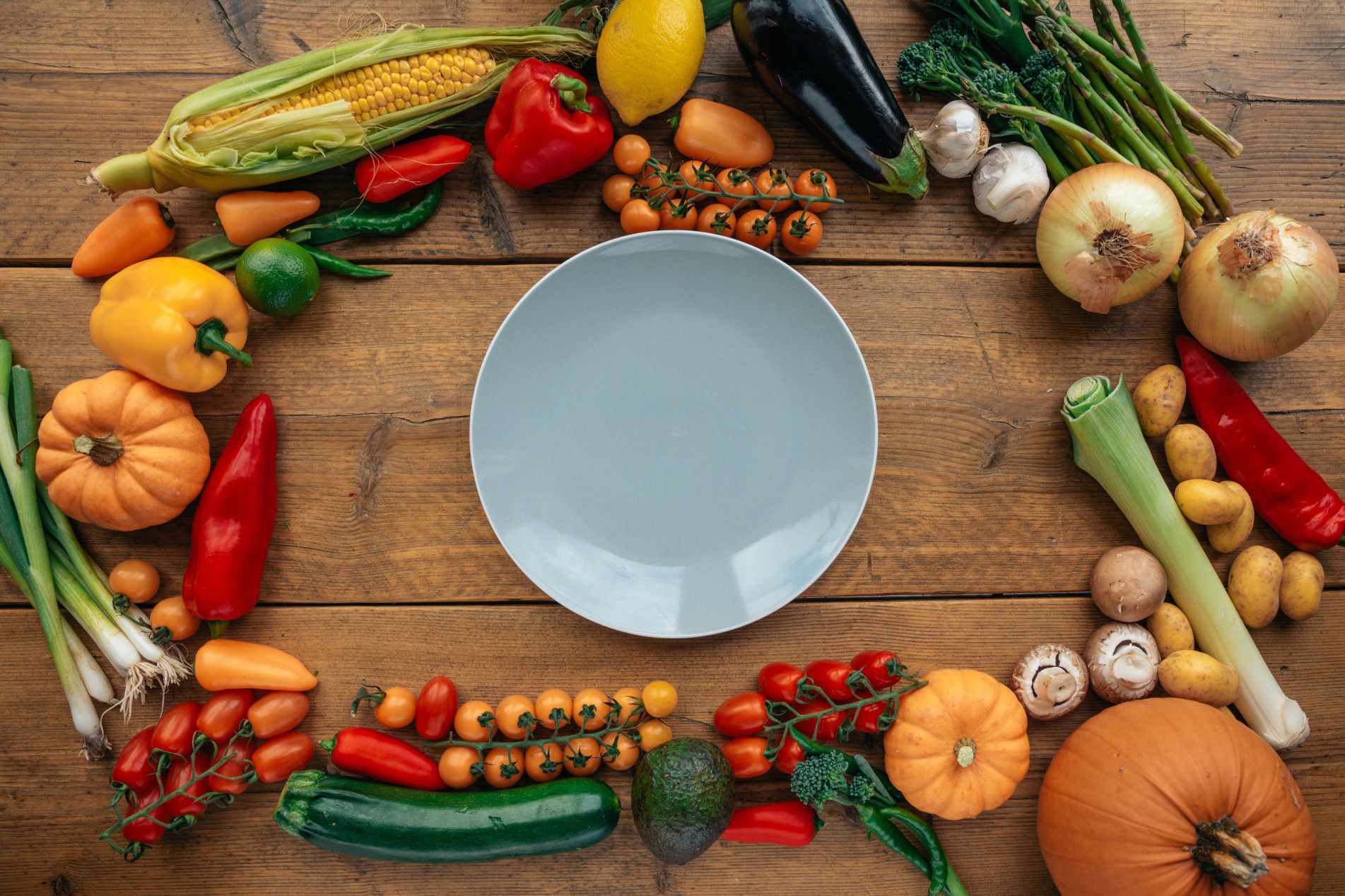 Prato vazio ao centro, cercado por uma variedade colorida de legumes e vegetais frescos sobre uma mesa de madeira.